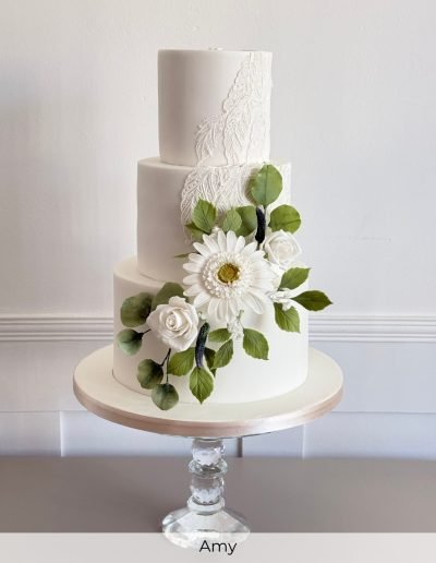 Elegant three-tier wedding cake with white lace detail and sugar daisies, designed by Anesha’s Art of Cakes