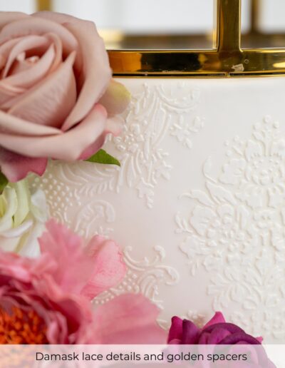 Ivory wedding cake with raised damask lace, pink flowers and polished gold geometric spacer.