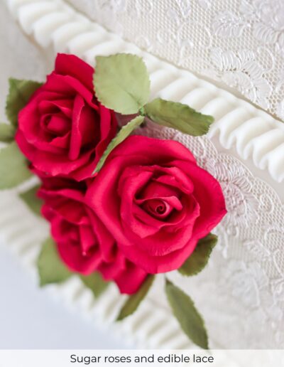 Close-up of deep red sugar roses, green leaves, ivory edible lace and piped wedding cake borders.