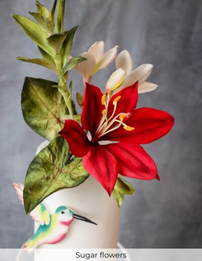 Red and ivory handcrafted sugar lilies with green leaves and a colourful hummingbird on an ivory wedding cake.